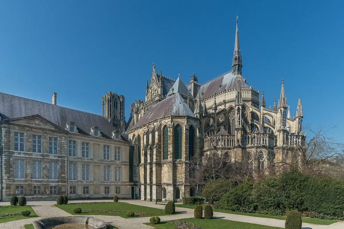 La Cattedrale di Reims vista da est con il cielo sereno, con archi rampanti, una guglia alta e un edificio classico in pietra sulla sinistra